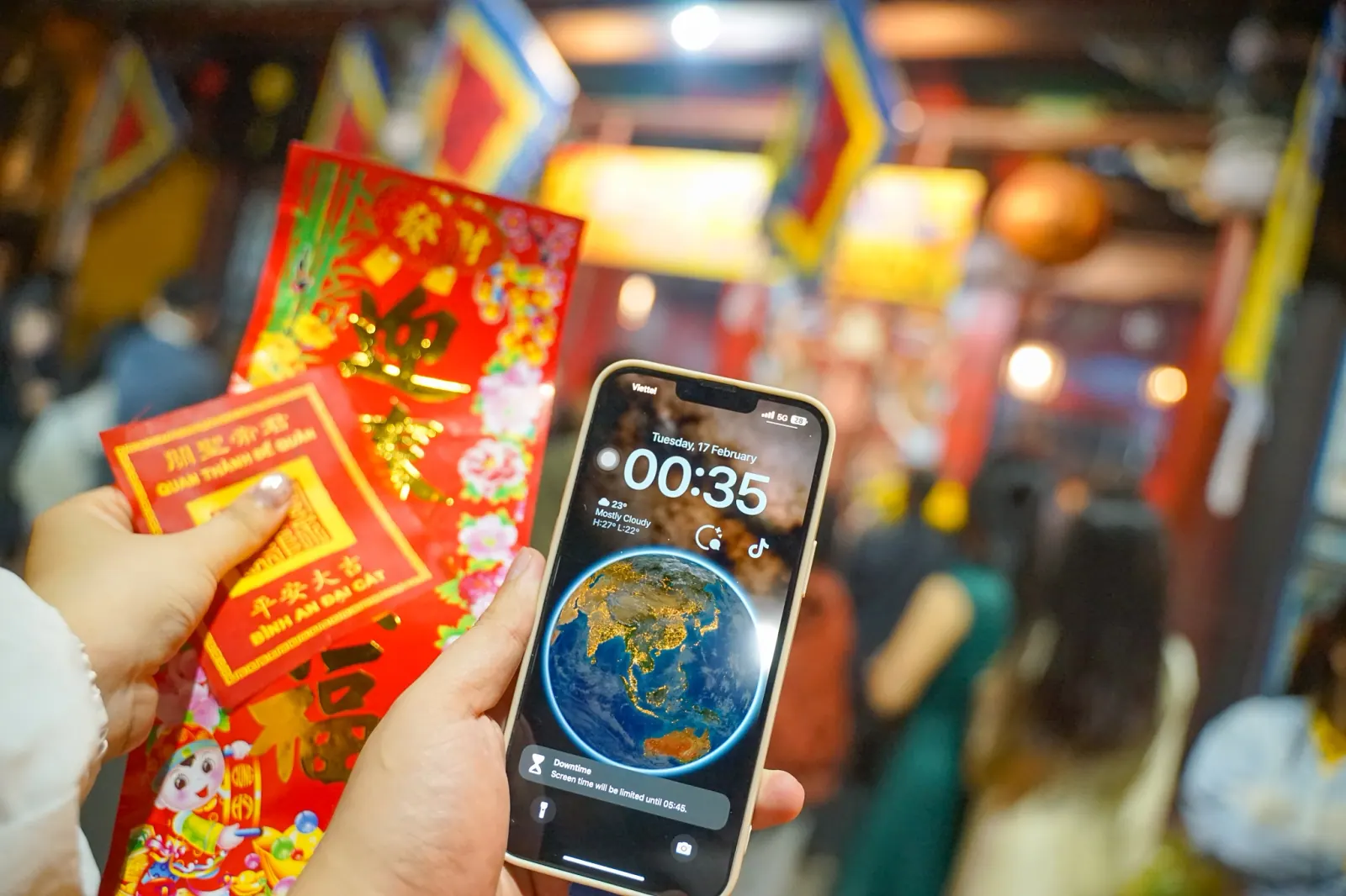Hand holding a red fortune envelope in the temple, phone screen reading 00:35 on the first morning of Tet